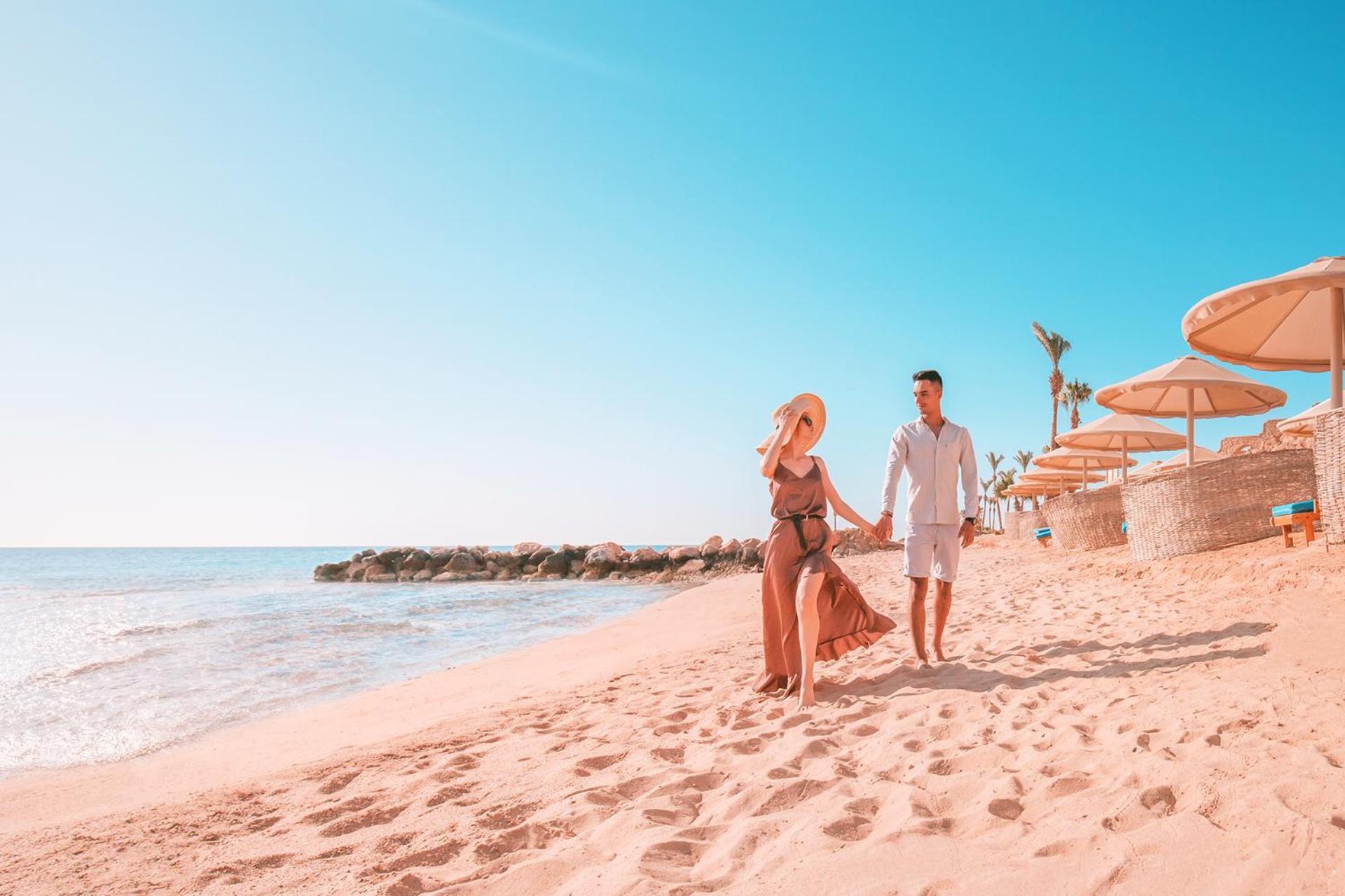 Pickalbatros Citadel Resort Sahl Hasheesh (Adults Only) Hurghada Exterior foto The photo depicts a couple walking hand in hand along a sandy beach. The woman is wearing a swimsuit with a flowing cover-up and a large sun hat, while the man is dressed in light shorts and a short-sleeve shirt. The beach is bathed in sunlight, and 