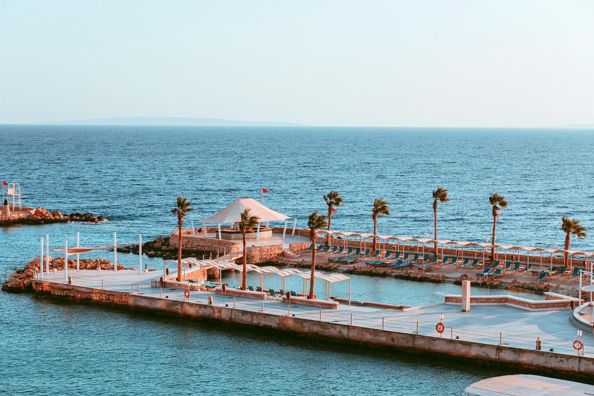 Pickalbatros Citadel Resort Sahl Hasheesh (Adults Only) Hurghada Exterior foto The photo shows a serene coastal scene with a clear blue sea and a light sky. In the foreground, there's a pier with several palm trees lining it. A white structure, possibly a pavilion, is situated near the shoreline. Additionally, there are sun lou