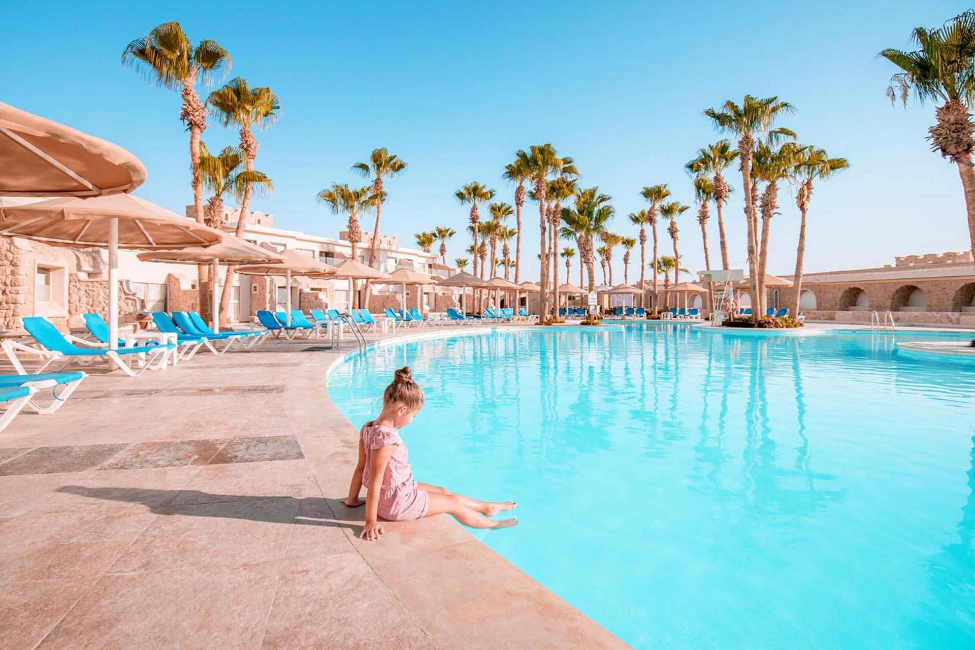 Pickalbatros Citadel Resort Sahl Hasheesh (Adults Only) Hurghada Exterior foto The photo shows a bright and inviting poolside scene. In the foreground, a young girl is sitting on the edge of a large, clear blue swimming pool, dipping her toes into the water. Surrounding the pool are tall palm trees and rows of lounge chairs wit