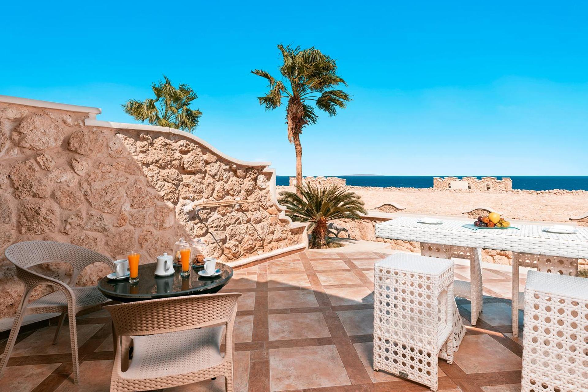 Pickalbatros Citadel Resort Sahl Hasheesh (Adults Only) Hurghada Exterior foto The photo shows a beautiful outdoor patio area with a view of the ocean. There are palm trees in the background, and the sky is clear and sunny. The seating includes a circular table with several chairs arranged around it. On the table, there are pla