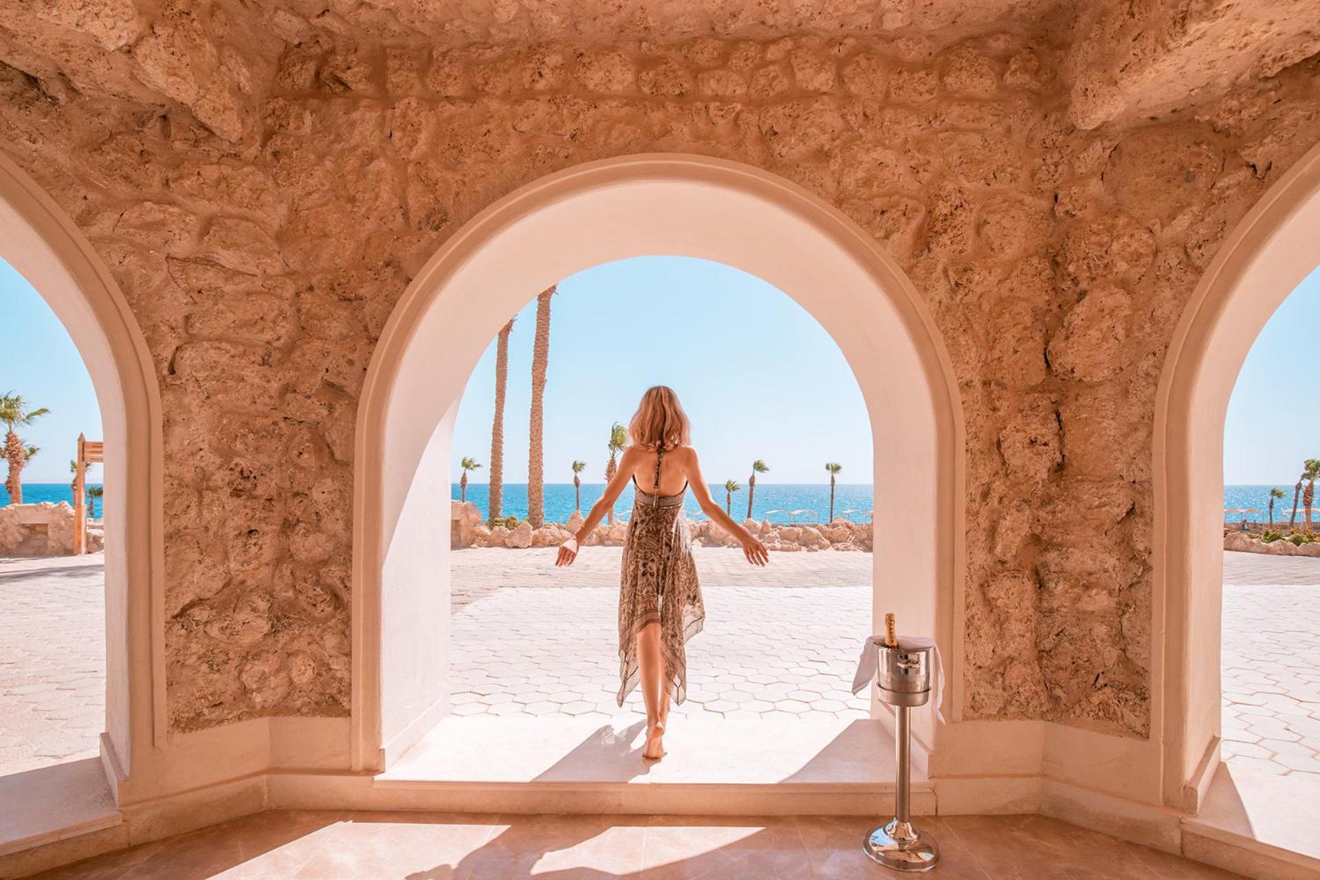 Pickalbatros Citadel Resort Sahl Hasheesh (Adults Only) Hurghada Exterior foto The photo depicts a woman standing at an archway that opens up to a scenic view. She is wearing a flowing, patterned dress and appears to be stepping outside. In the background, the vibrant blue sea can be seen, along with palm trees and a clear sky 