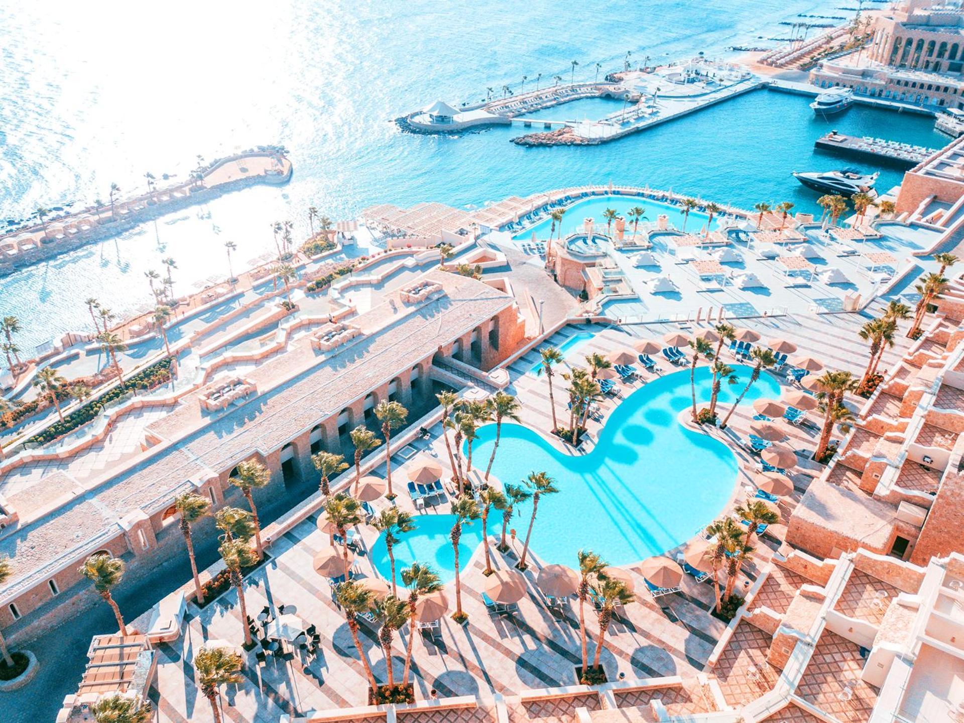 Pickalbatros Citadel Resort Sahl Hasheesh (Adults Only) Hurghada Exterior foto The photo shows a luxurious resort with a large, shaped swimming pool surrounded by palm trees and lounge chairs. The pool has a unique curve design, enhancing the overall aesthetic. In the background, there's a beautiful waterfront view with boats a